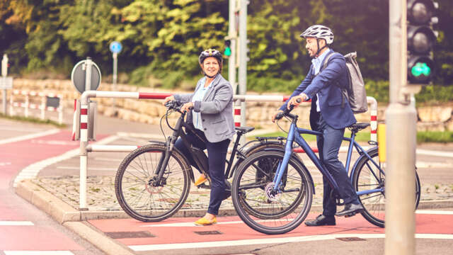 zwei Radfahrer:innen stehen nebeneinander, leicht versetzt, vor einer Ampel auf einem rot markierten Fahrradweg