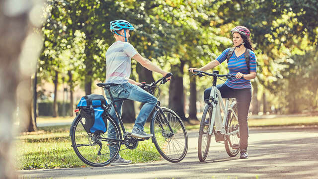 zwei RadfahrerInnen treffen sich auf einem Weg mit Bäumen im Hintergrund und halten an um sich zu unterhalten.